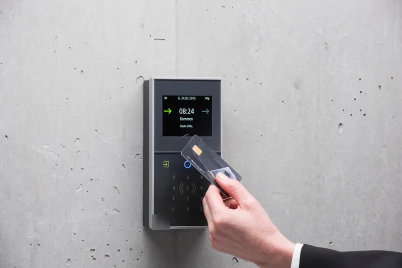 A hand presenting a card to a wall-mounted reader with a small screen and keypad