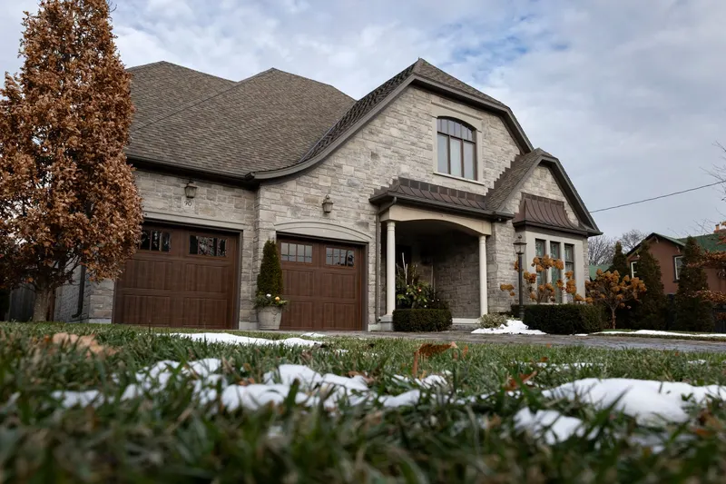 Two brown carriage-style garage doors on a stone house, with a light dusting of snow on the lawn