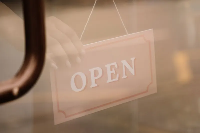 A hand turning a hanging Open sign on a glass shop door beside a curved metal pull handle