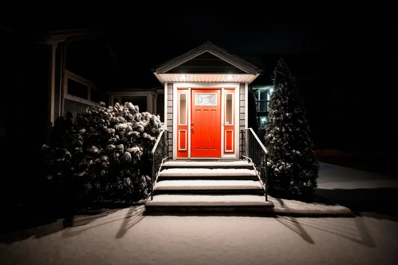 A porch light glowing over a red front door on a snowy night